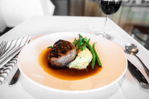 A plated steak with mashed potato and green beans beside a glass of red wine on white linen